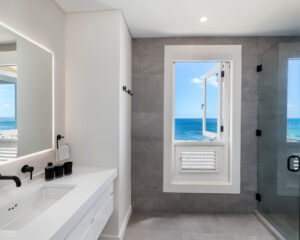 bathroom of a luxury villa with white fixtures and a large mirror on the left, a white window with a view of the ocean in front and a glass shower on the right.