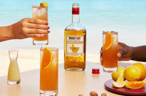 three glasses of rum punch on a table, two held in hands with a bottle of mount gay rum in the background.